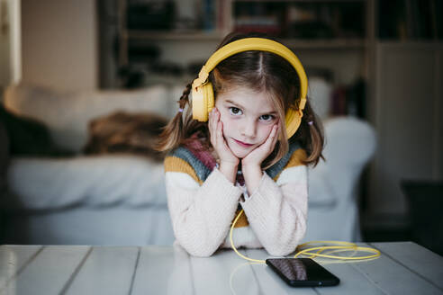 Mädchen mit Kopfhörern lehnt sich zu Hause mit dem Kopf in den Händen an einen Tisch - EBBF01135