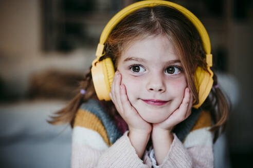 Cute girl with headphones and head in hands looking away while standing at home - EBBF01136