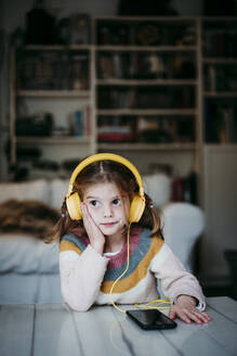 Mädchen mit Kopfhörern, das wegschaut, während es sich zu Hause auf einen Tisch stützt - EBBF01138