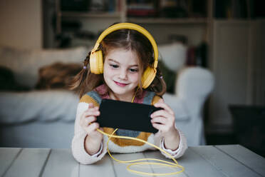 Lächelndes Mädchen mit Kopfhörern, das ein Mobiltelefon benutzt, während es zu Hause steht - EBBF01139