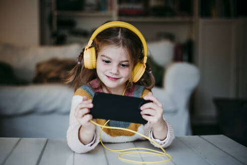 Smiling girl wearing headphones using mobile phone while standing at home - EBBF01139