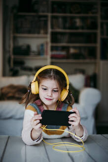 Girl wearing headphones using mobile phone while standing at home - EBBF01140