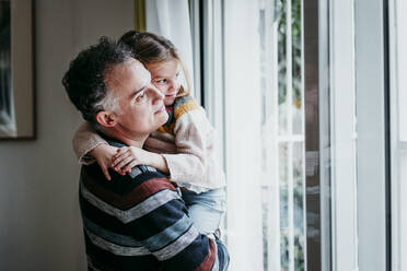 Father carrying daughter while standing by window at home - EBBF01141