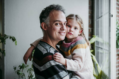 Lächelnder Vater trägt seine Tochter am Fenster zu Hause, lizenzfreies Stockfoto