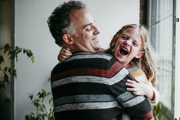 Lächelnder Vater mit verspielter Tochter am Fenster stehend zu Hause - EBBF01145