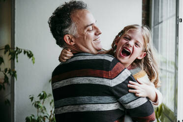 Smiling father carrying playful daughter while standing by window at home - EBBF01145