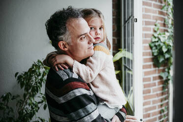 Father with daughter looking through window while standing at home - EBBF01147