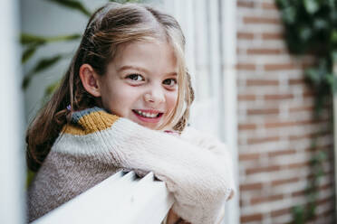 Smiling girl leaning on window while standing at home - EBBF01148