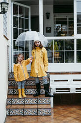 Sisters wearing raincoat holding umbrella while standing on staircase - EBBF01150