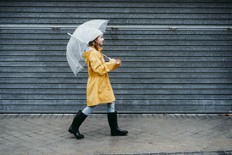 Girl wearing raincoat and jump boot holding umbrella while walking on sidewalk stock photo