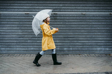 Mädchen in Regenmantel und Springerstiefel hält Regenschirm beim Gehen auf dem Bürgersteig - EBBF01154