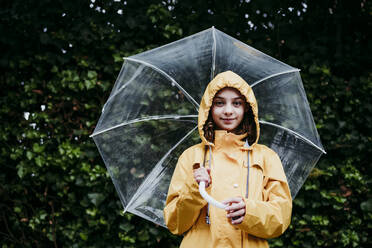 Mädchen mit Regenmantel, das einen Regenschirm hält, während es an einer Laubwand steht - EBBF01157