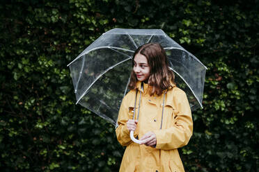 Girl wearing raincoat looking away while standing against leaf wall - EBBF01158