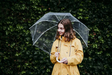 Mädchen in Regenmantel schaut weg, während sie gegen eine Laubwand steht - EBBF01158
