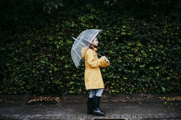 Mädchen mit Regenmantel und Regenschirm, das gegen eine Laubwand in der Stadt läuft - EBBF01159