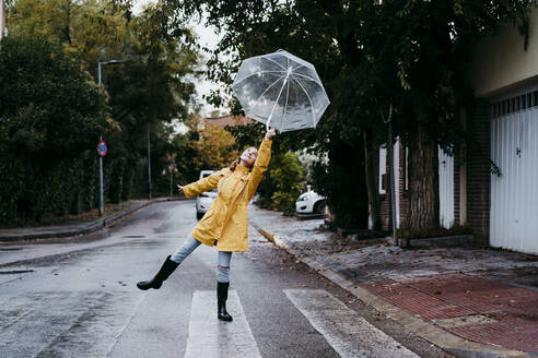 Girl wearing raincoat dancing with umbrella while standing on road in city - EBBF01160