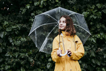 Girl in raincoat with umbrella looking away while standing against leaf wall - EBBF01162