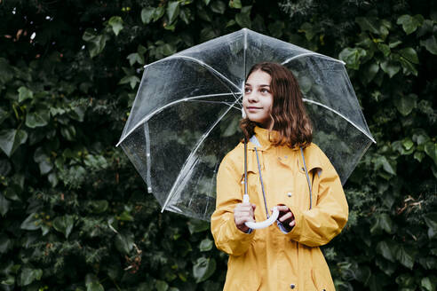 Girl in raincoat with umbrella looking away while standing against leaf wall - EBBF01162