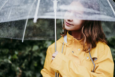 Girl in raincoat looking away while standing under umbrella outdoors - EBBF01163