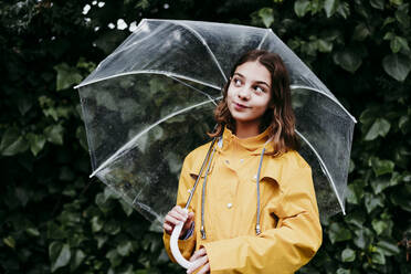 Smiling girl in raincoat with umbrella looking away while standing against leaf wall - EBBF01166
