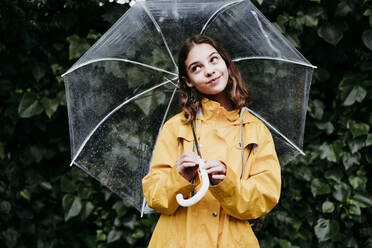 Thoughtful girl smiling while holding umbrella against leaf wall - EBBF01167