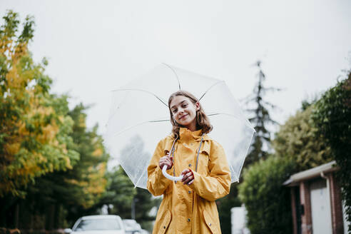 Smiling girl in raincoat holding umbrella while standing in city - EBBF01169