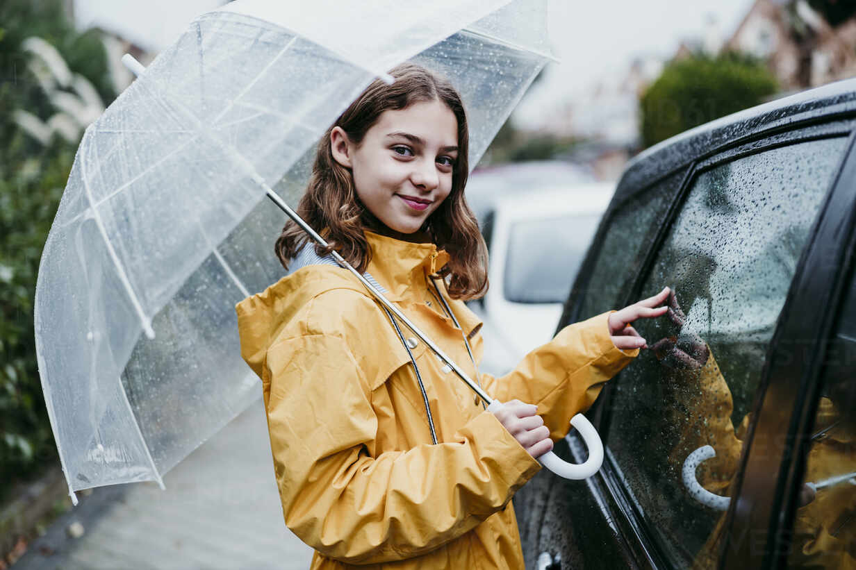 Lächelndes Mädchen in Regenmantel mit Regenschirm in der Hand, während sie  in der Stadt am Auto steht, lizenzfreies Stockfoto