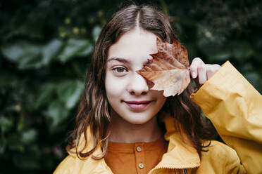 Smiling girl in raincoat hiding eye with dry ivy leaf while standing against leaf wall - EBBF01173