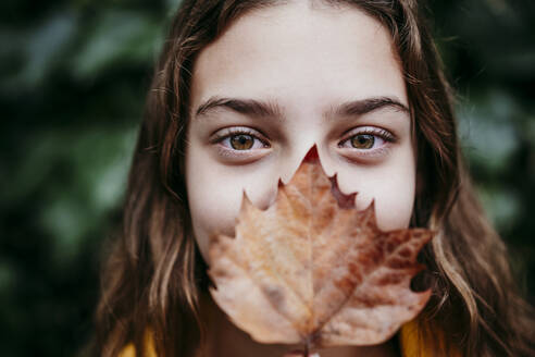 Girl in raincoat hiding face with dry ivy leaf while standing against leaf wall - EBBF01174