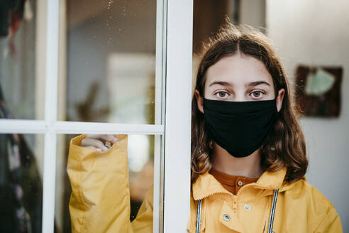 Girl wearing raincoat and face mask standing by door at home - EBBF01176