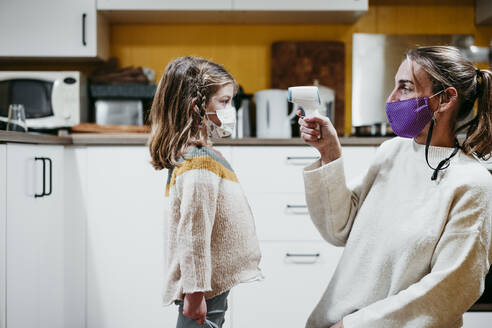 Mother wearing face mask checking temperature of daughter while sitting at home - EBBF01181