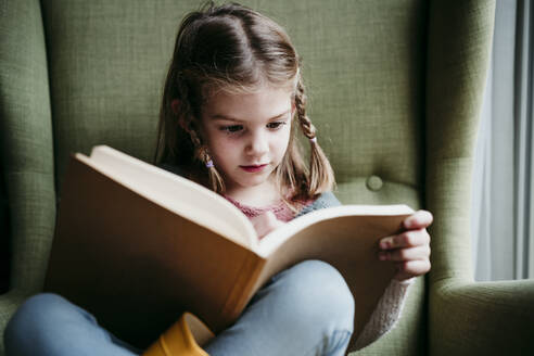 Girl reading book while sitting on chair at home - EBBF01185