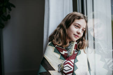 Girl covered in blanket smiling while sitting by window at home - EBBF01191