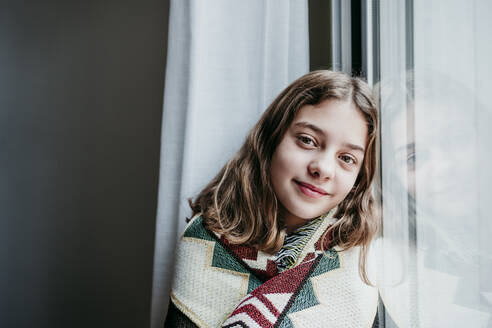 Smiling girl covered in blanket sitting by window at home - EBBF01192