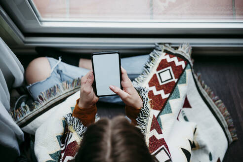 Mädchen mit Decke, das ein Mobiltelefon benutzt, während es zu Hause am Fenster sitzt - EBBF01193