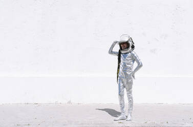 Smiling boy in astronaut costume saluting while standing against wall - JCMF01589