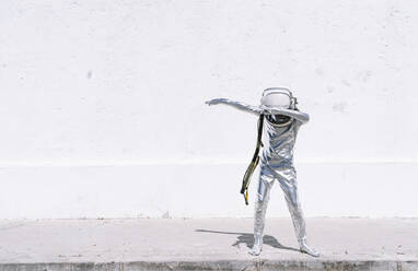 Boy in astronaut costume dabbing movement while standing against wall - JCMF01591