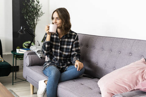 Frau trinkt Kaffee, während sie zu Hause auf dem Sofa sitzt, lizenzfreies Stockfoto