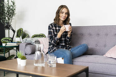 Young woman drinking coffee while sitting on sofa at home - GIOF09332