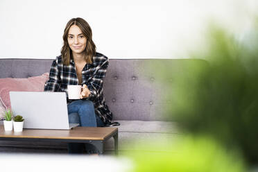 Junge Frau trinkt Kaffee und benutzt einen Laptop zu Hause - GIOF09333