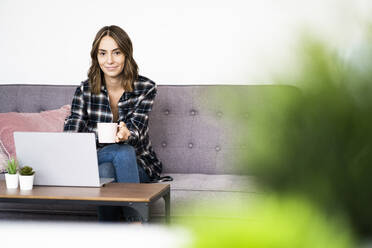 Young woman drinking coffee while using laptop at home - GIOF09333