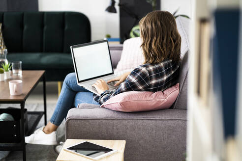 Junge Frau arbeitet am Laptop, während sie zu Hause auf dem Sofa sitzt - GIOF09337