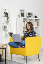 Junge Frau benutzt einen Laptop, während sie zu Hause auf dem Sofa sitzt - GIOF09339