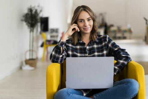 Lächelnde junge Frau mit dem Kopf in den Händen bei der Arbeit am Laptop, während sie zu Hause auf dem Sofa sitzt - GIOF09341
