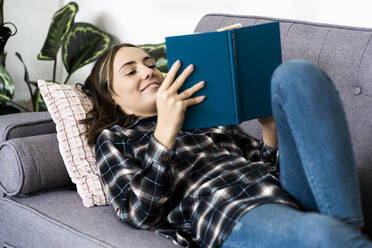 Smiling woman reading book while lying on sofa at home - GIOF09343