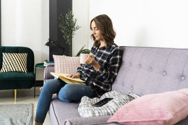 Young woman with coffee cup reading book while sitting on sofa at home - GIOF09344