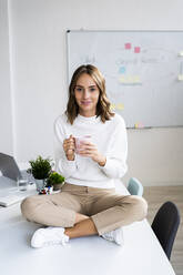 Young businesswoman drinking coffee while sitting on table at office - GIOF09375