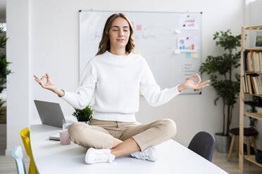 Junge Geschäftsfrau übt Yoga, während sie auf dem Tisch im Büro sitzt - GIOF09376