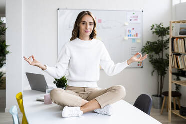 Junge Geschäftsfrau macht Yoga, während sie auf dem Tisch im Büro sitzt - GIOF09377