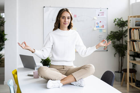 Junge Geschäftsfrau macht Yoga, während sie auf dem Tisch im Büro sitzt - GIOF09377